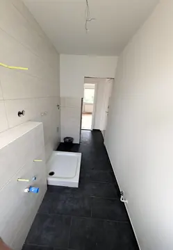 Langes, schmales Bad mit hellen Wandfliesen und dunklem Boden, vorn die Duschwanne
